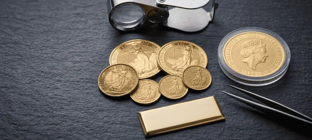 Gold Britannia coins with a loupe, a capsuled coin and a gold bar on slate