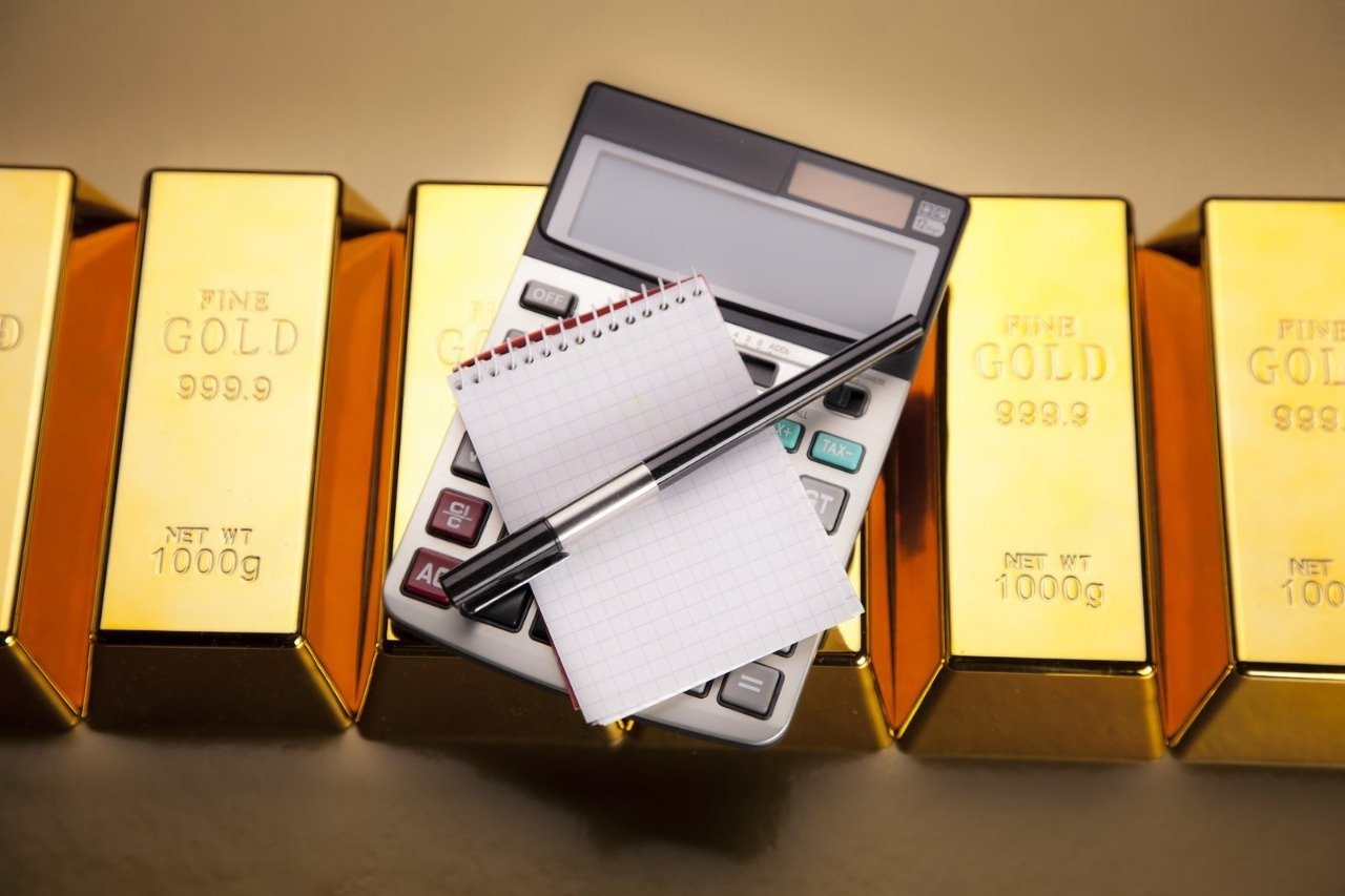 A row of gleaming 1000g Fine Gold Bullion bars with a calculator, spiral notepad, and pen resting on top, representing the strategic financial planning and Precious Metal Investment strategies detailed in our comprehensive Gold Investment Guide for those exploring the Best Way to Buy Gold.