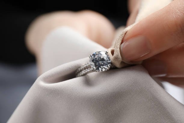 A close-up view of a person using a soft beige polishing cloth to gently wipe down a sparkling diamond engagement ring, illustrating the fundamental steps of gold care and maintenance and proper jewelry care.