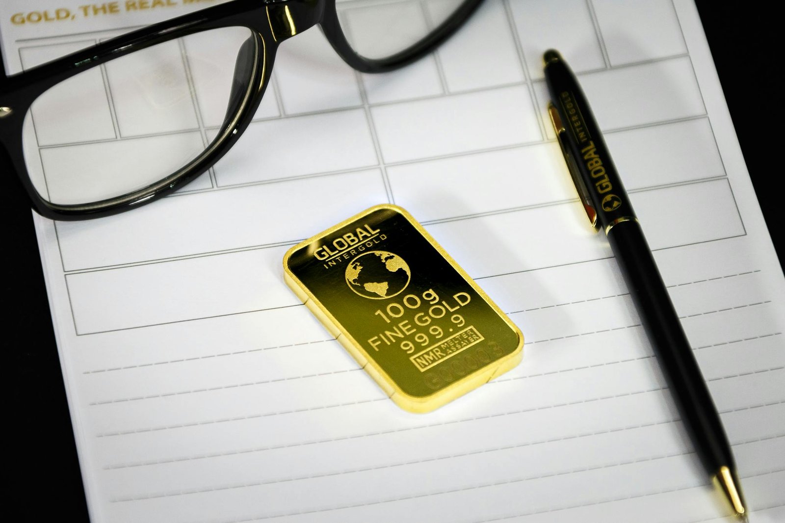 A 100g fine gold bullion bar stamped with official purity markings resting on a white lined paper document next to a pen and reading glasses, providing a clean visual example for a comprehensive gold hallmarks guide.