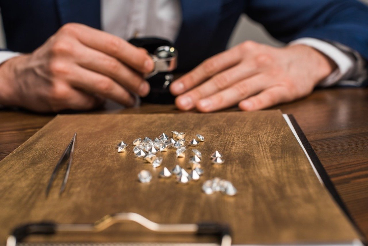 A close-up view of a cluster of loose brilliant-cut diamonds resting on a wooden clipboard alongside fine tweezers, while a jeweler uses a precision loupe in the blurred background, highlighting the selection process for Lab Diamonds Little Rock clients select for custom jewelry.
