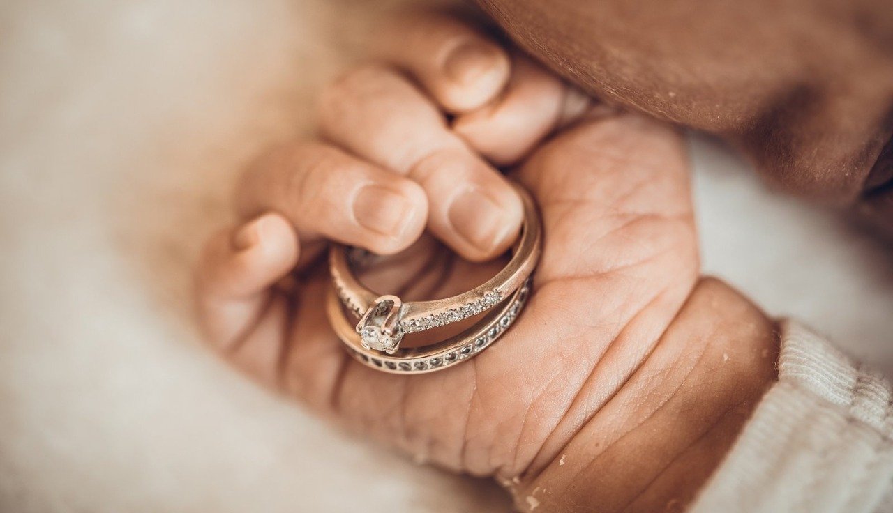 A close-up, tender photograph of a newborn baby's tiny hand resting on soft bedding while holding a delicate gold diamond ring set, highlighting the beautiful sentiment of gifting premium Newborn Jewelry Little Rock family heirlooms.